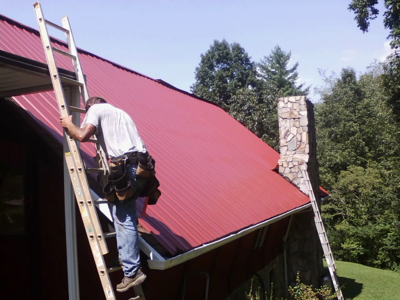 Roofer on a ladder beside a red metal roof and stone chimney — Soffit & Fascia Repair by Roofing Brothers in Bethune